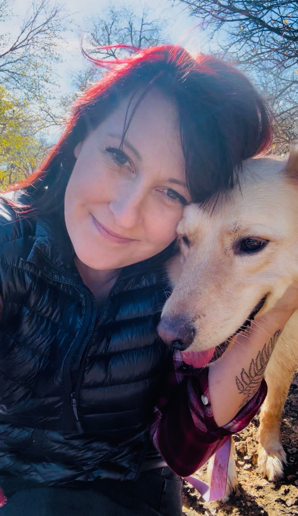 Woman with red-highlighted hair smiling cheek-to-cheek with a golden retriever wearing a bandana outdoors.