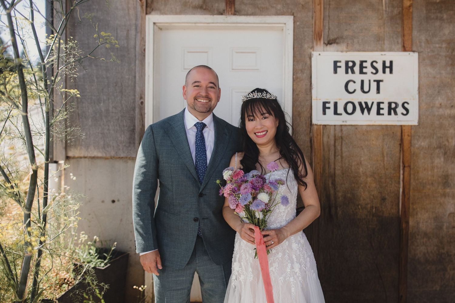 Bride and groom smiling together outside rustic wooden building with Fresh Cut Flowers sign, bride holding purple bouquet.