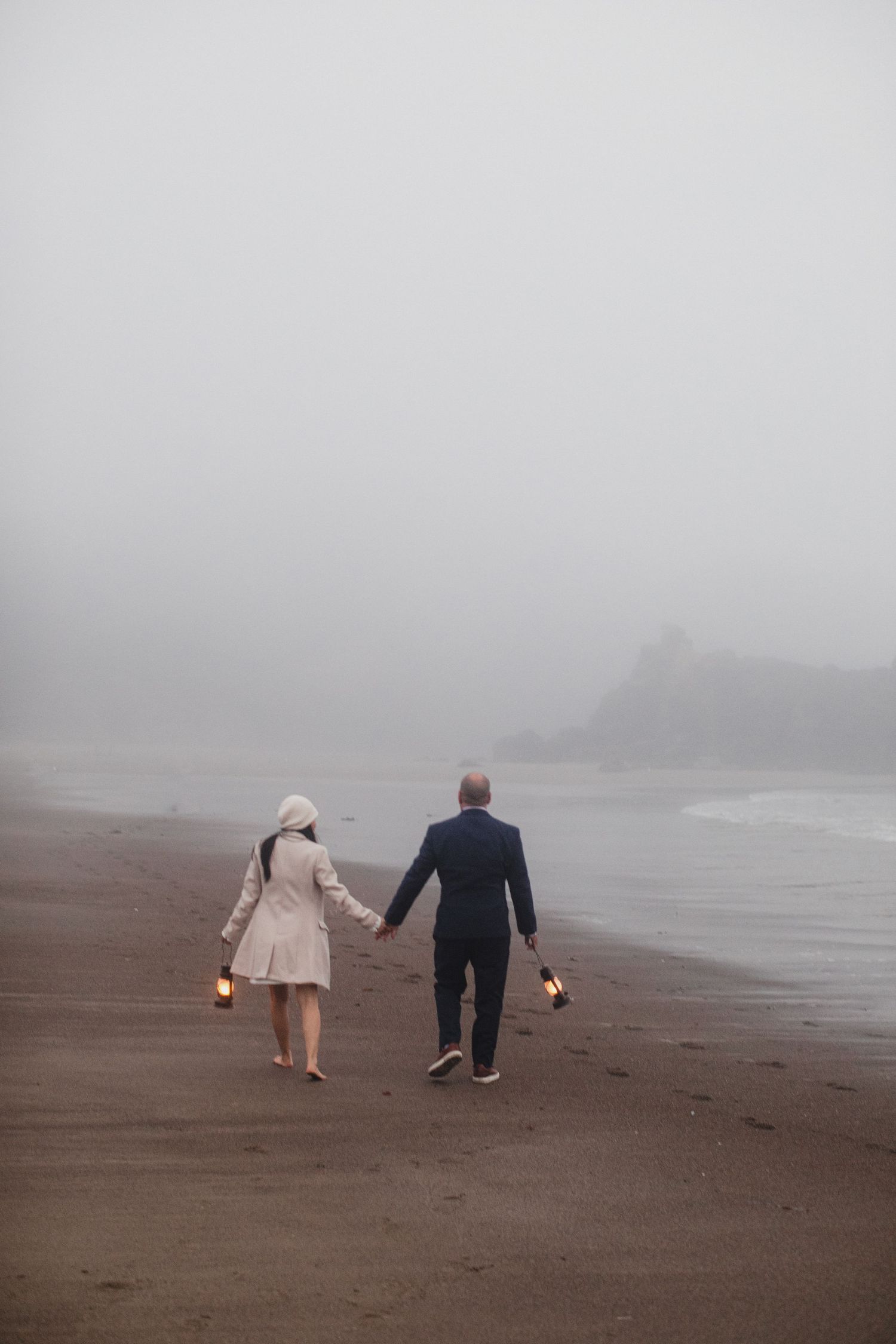 Couple holding lanterns walking hand in hand on a foggy misty beach at dusk.