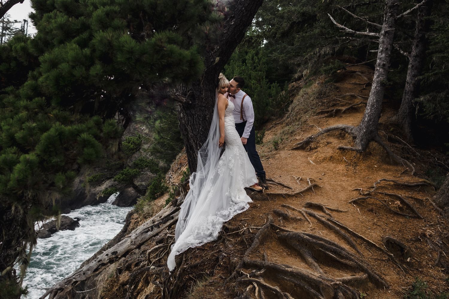 A wedding couple stands on a rocky cliffside path overlooking the churning ocean below.