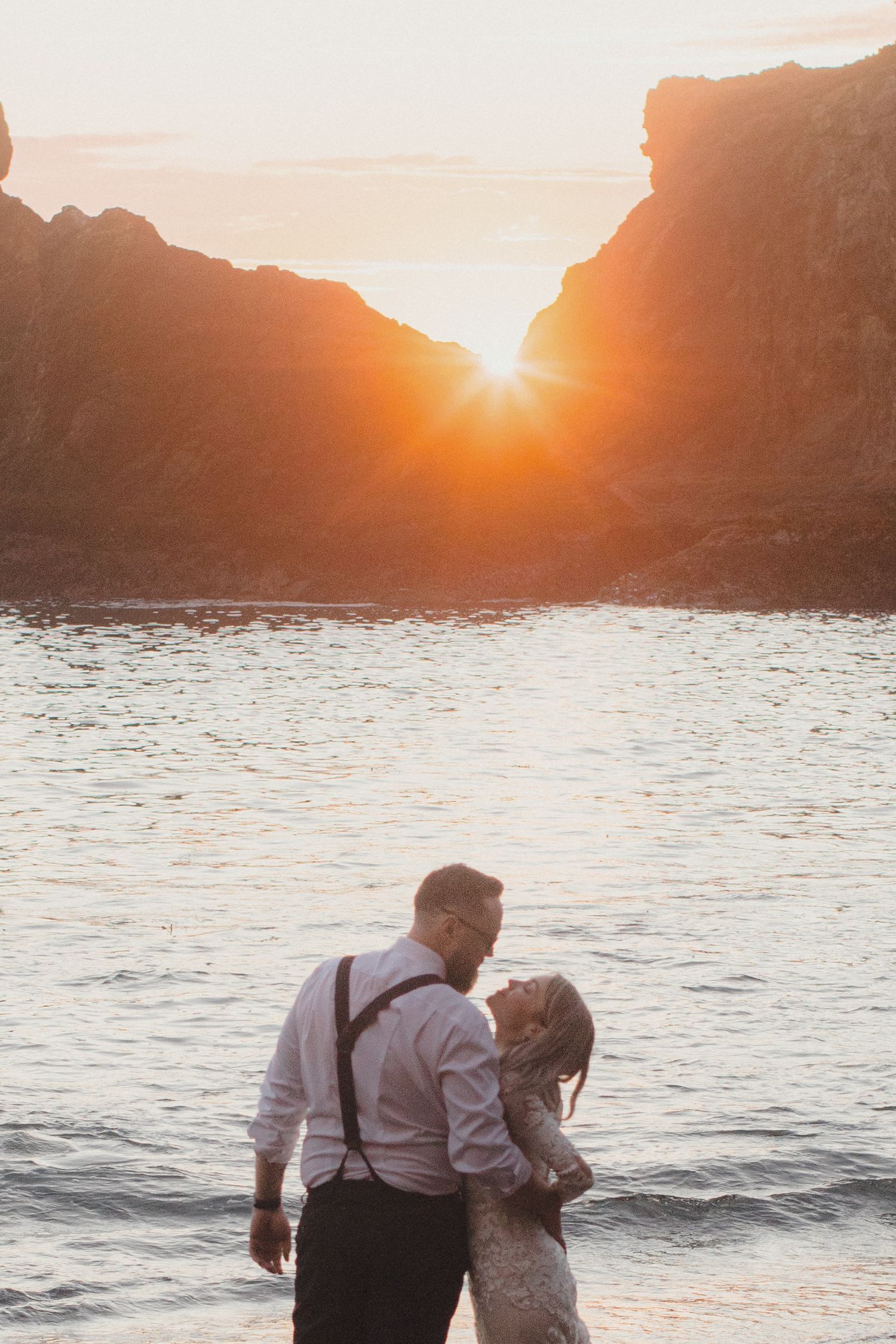 Couple kissing on a beach at sunset between two rocky cliffs.