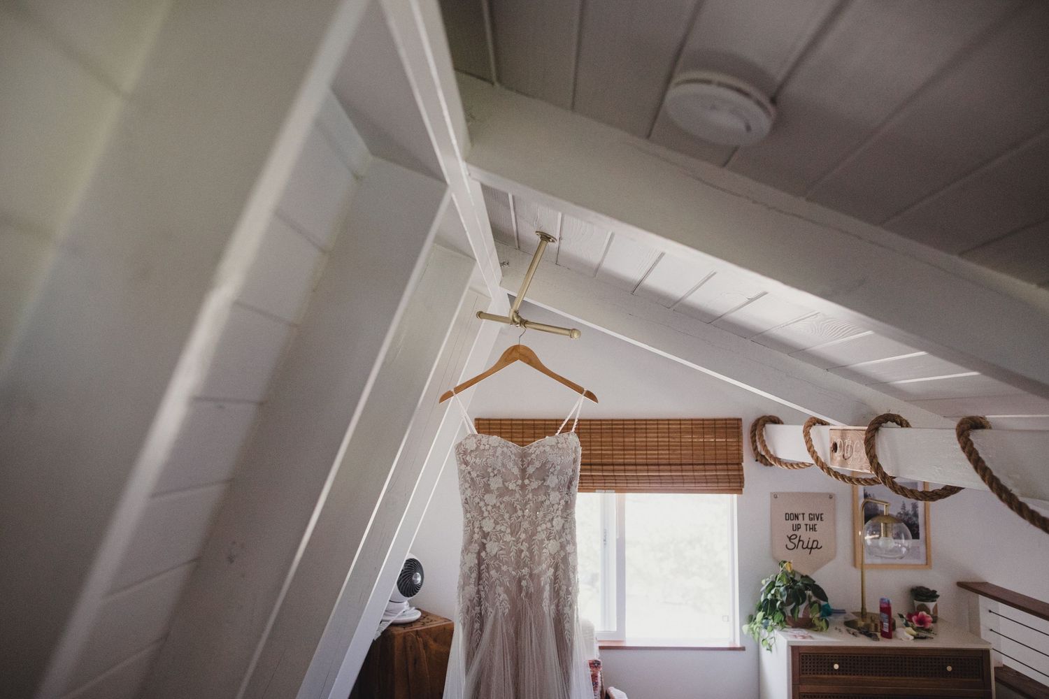 Wedding dress hanging on a wooden hanger in a rustic attic room with white slanted ceiling.