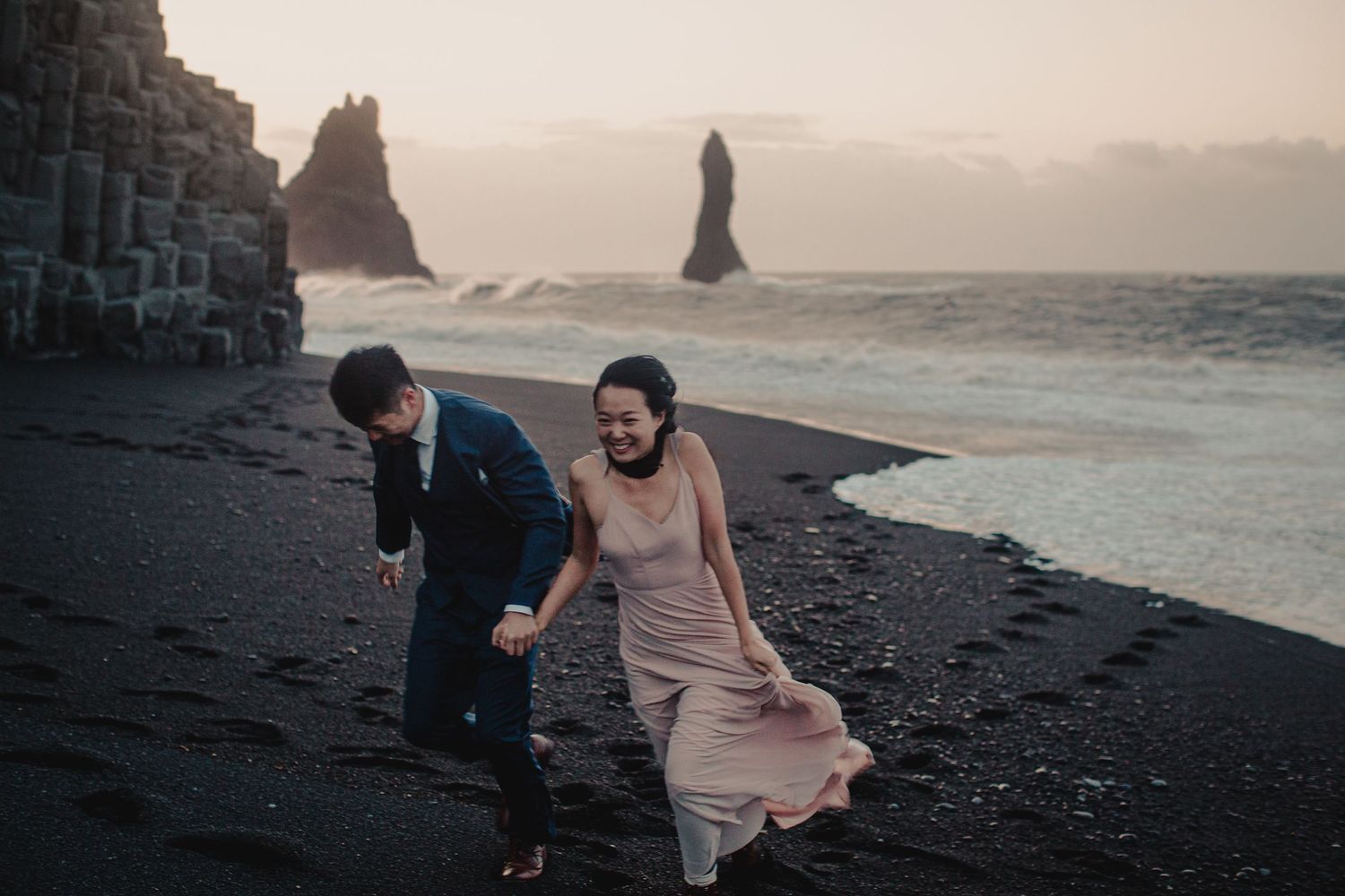 Two people walking together on a dramatic black sand beach with rock formations and misty coastline at sunset.