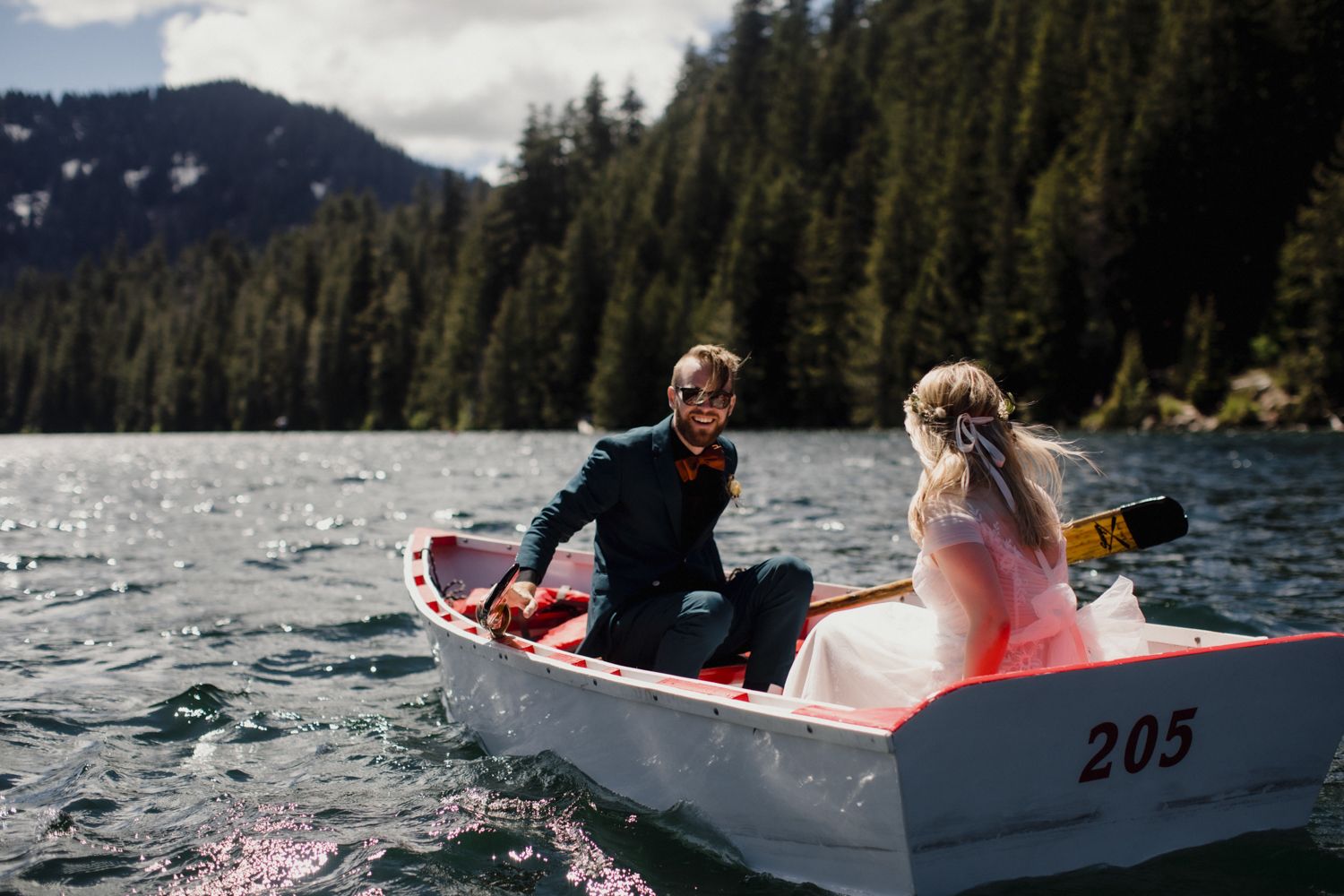 Couple rowing small boat on mountain lake with evergreen forest backdrop.