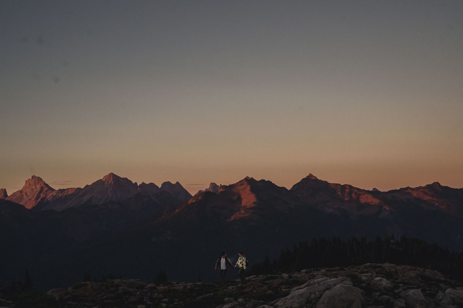 Jagged mountain peaks glow with soft orange alpenglow at dusk while two hikers stand on a rocky ridge in the foreground.