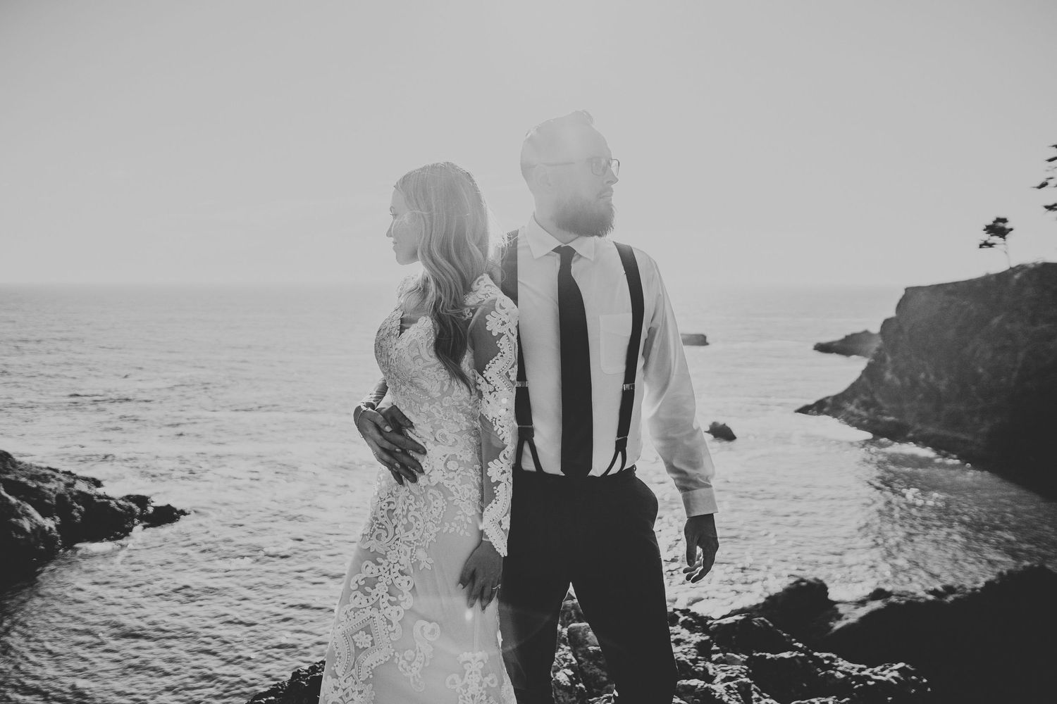 A bride and groom stand back-to-back in black and white photo overlooking dramatic ocean cliffs.