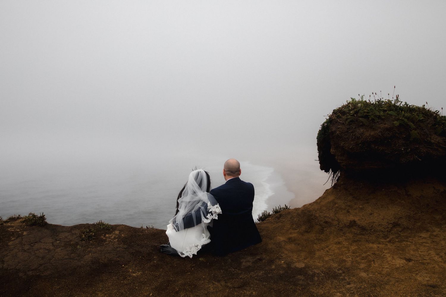 Bride and groom sitting together on a misty coastal cliff on their wedding day.