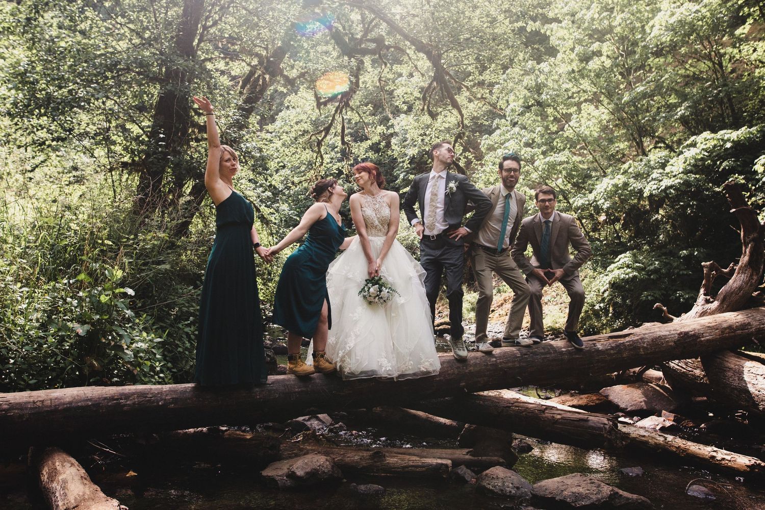 Wedding party stands on fallen log over creek in lush forest at Silver Falls State Park on their elopement day