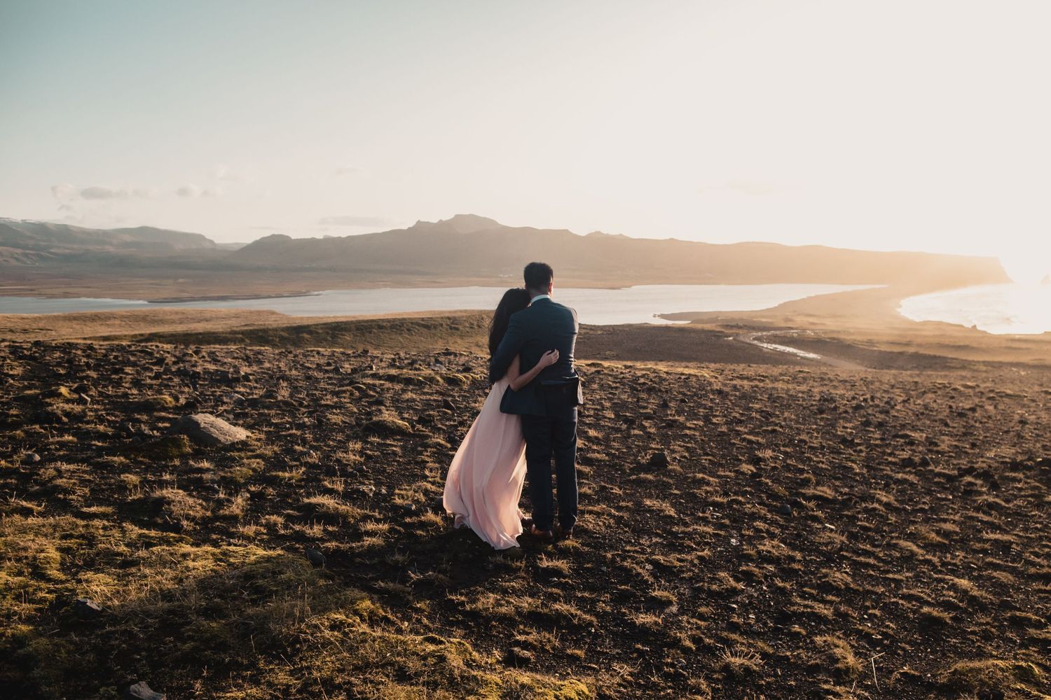 A couple embraces during sunset on a rugged coastal clifftop with mountains and water in the background.