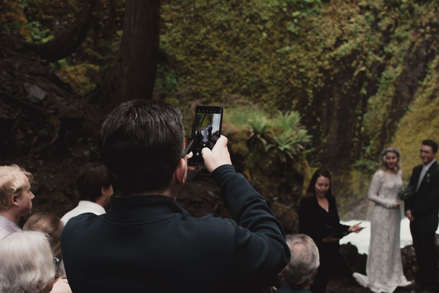 Person taking photo with smartphone at outdoor gathering in wooded setting.