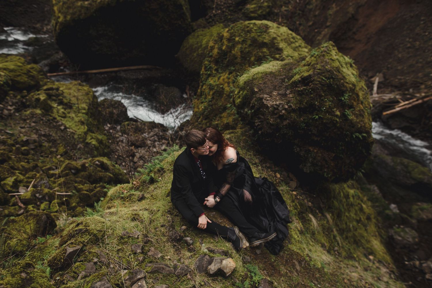 Couple in black clothing sitting intimately on mossy rocks beside a forest stream in a lush, green wilderness setting.