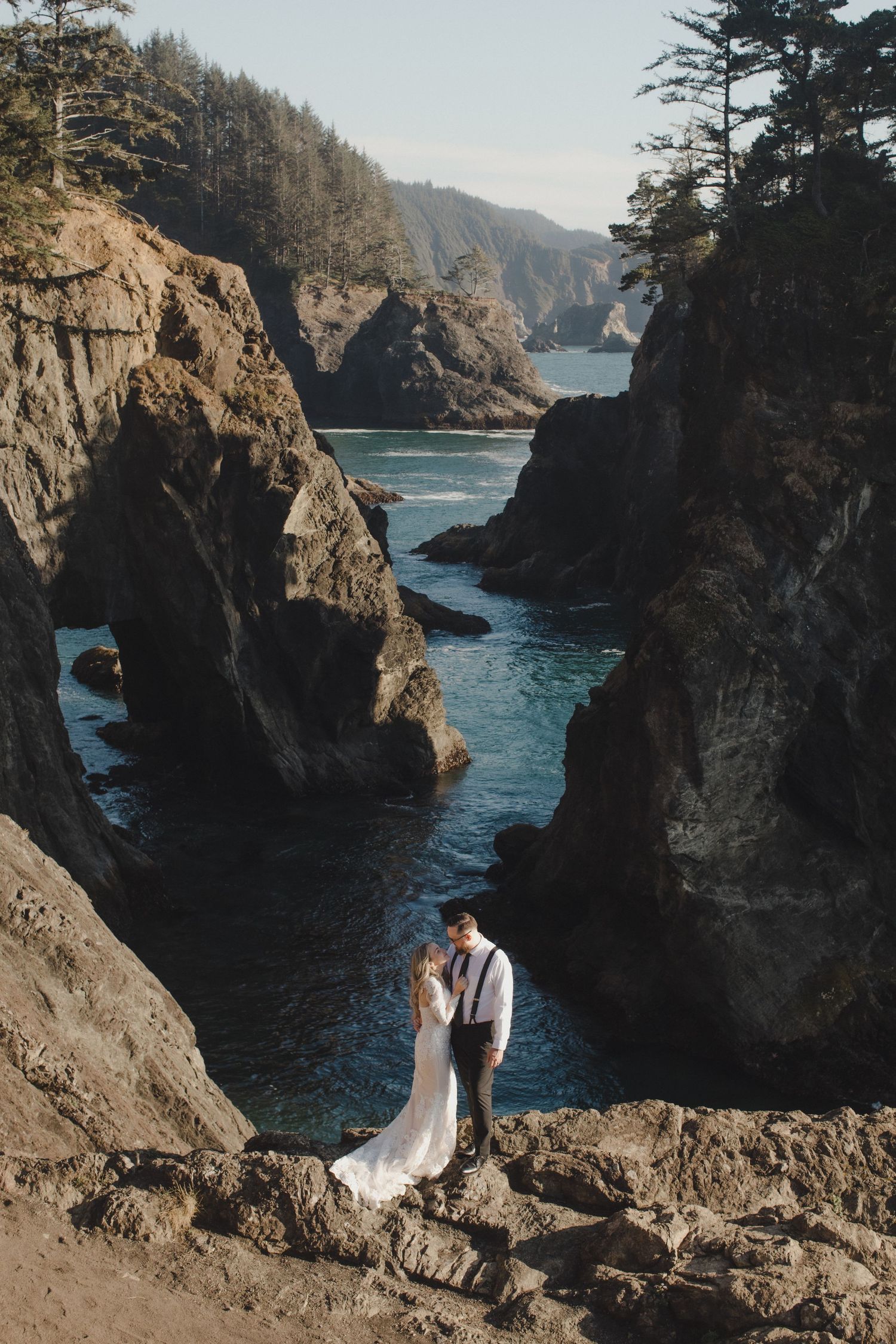 Bride and groom embrace on rocky coastal cliffs with turquoise ocean waters and sea stacks in background.
