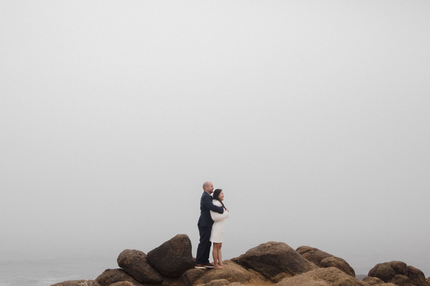 Couple embracing on rocky cliffs overlooking the ocean on a foggy wedding day.