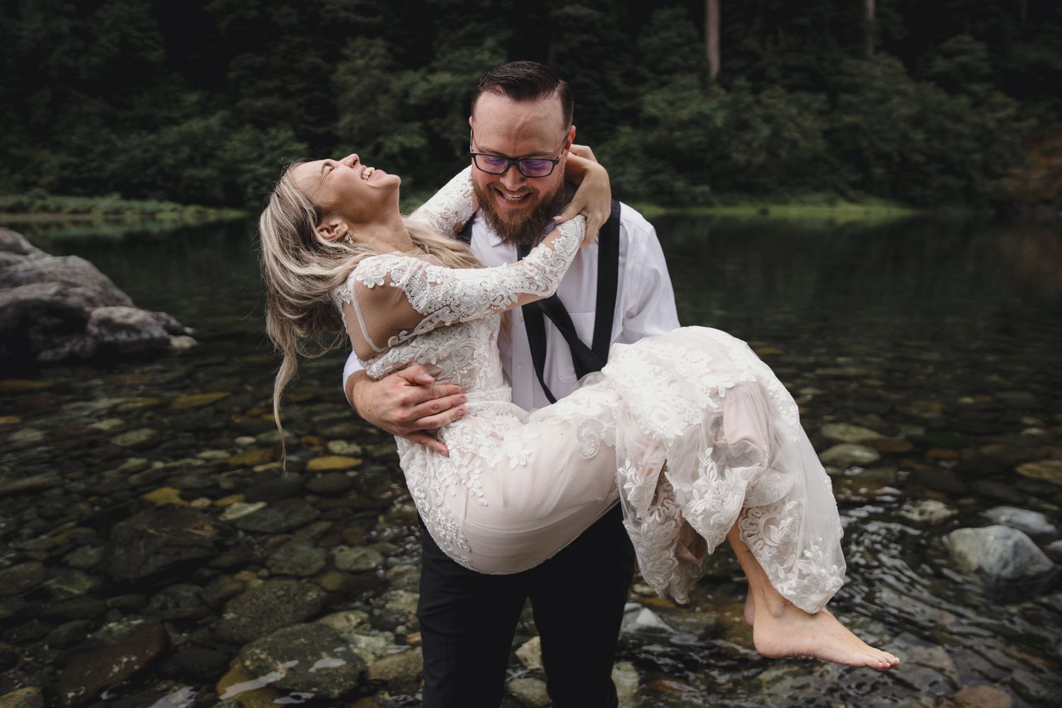 Groom lifting bride in white lace dress by a rocky river, laughing joyfully during outdoor wedding photo session.