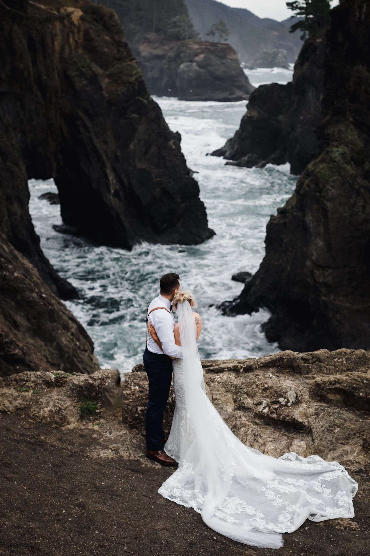 A couple stands at the edge of dramatic coastal cliffs while waves crash through the rocky gorge below.