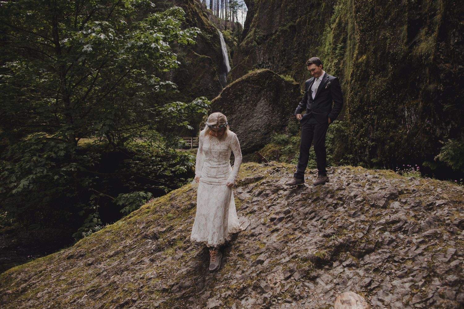 Two people stand on rocky ledge with waterfall visible through forest trees.