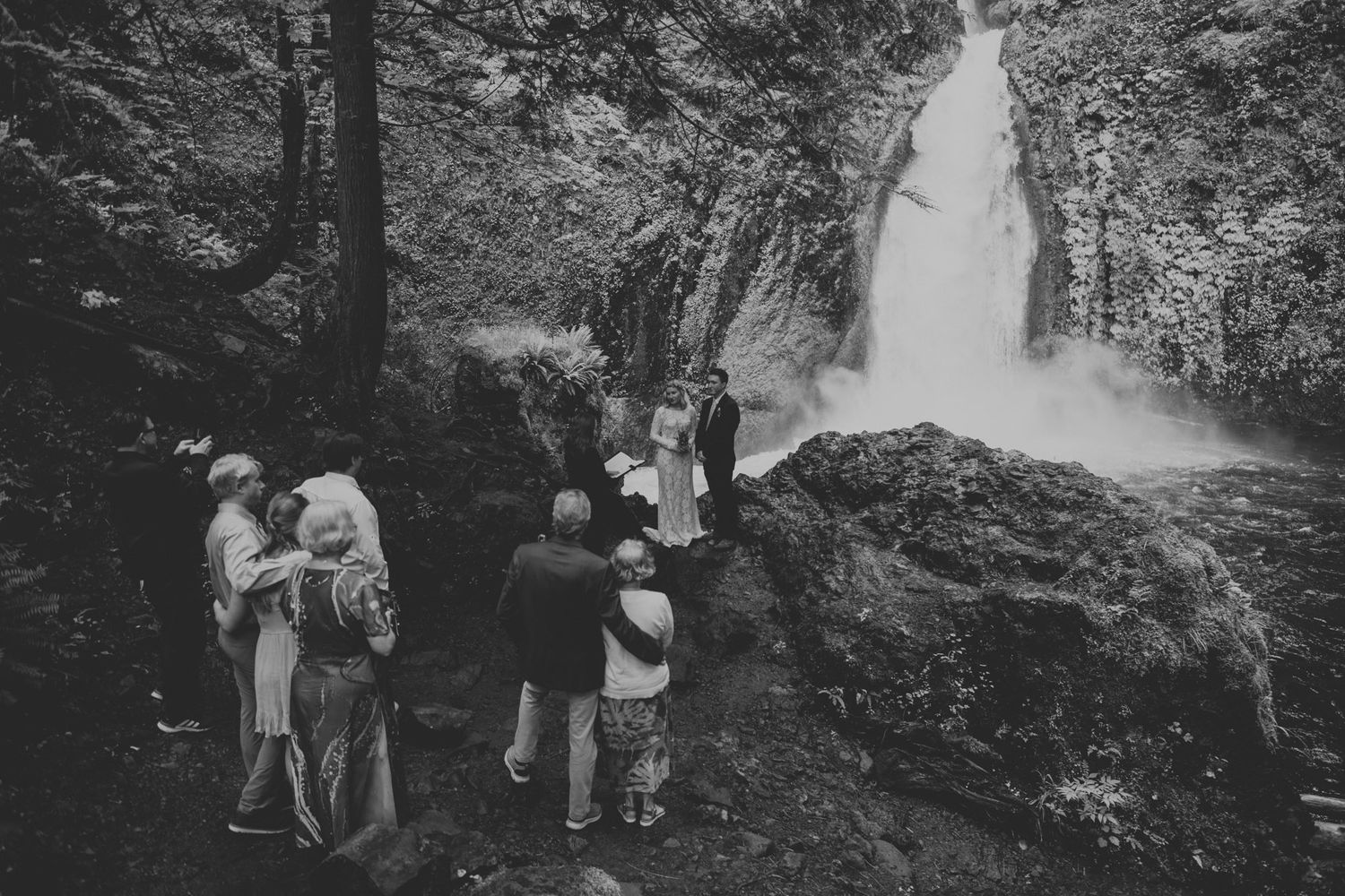 Small group gathers near dramatic waterfall in black and white forest scene.