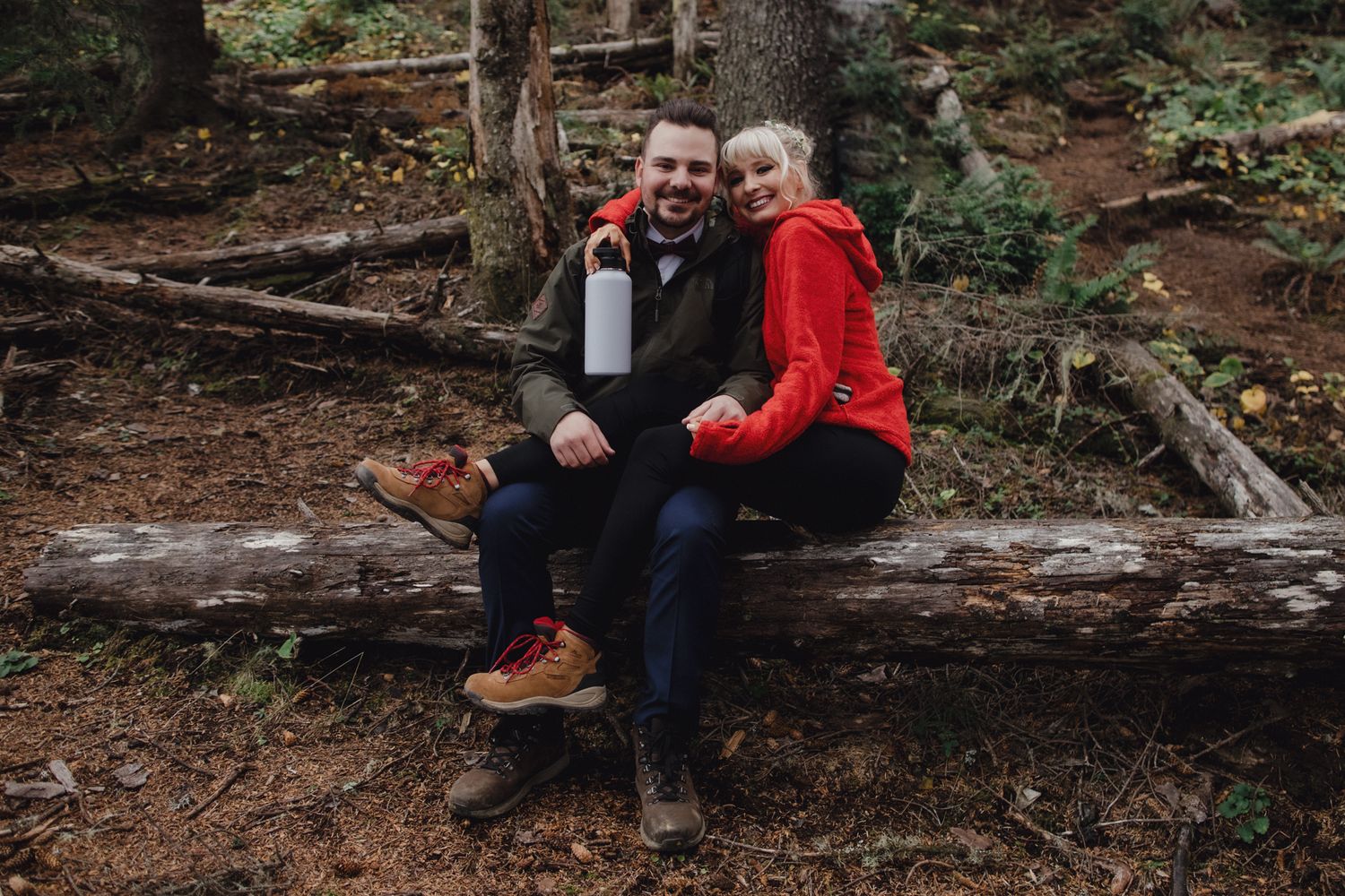 A couple shares a cheerful moment while sitting on a fallen log in a forest setting.