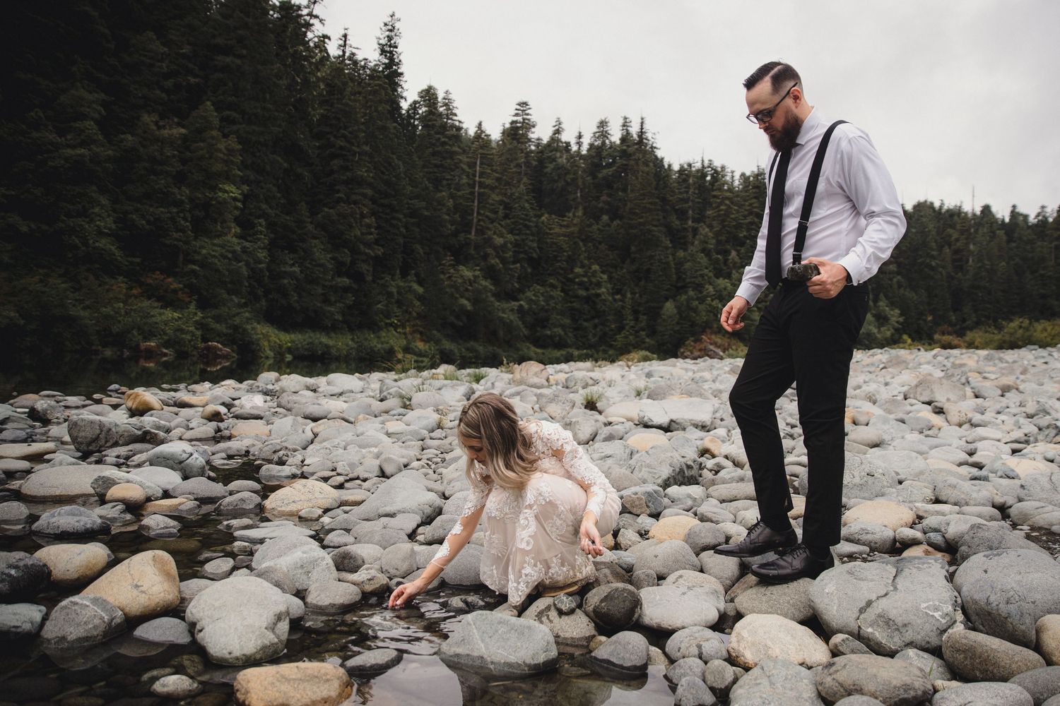 Bride in lace dress touches rocks by river as groom in suspenders watches during outdoor elopement.