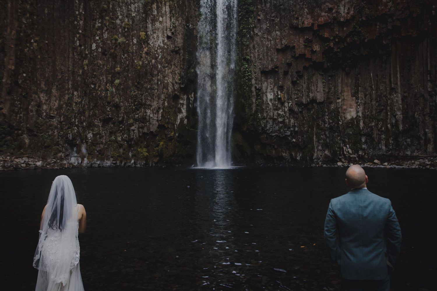 Bride and groom stand before a stunning waterfall cascading into a dark pool.