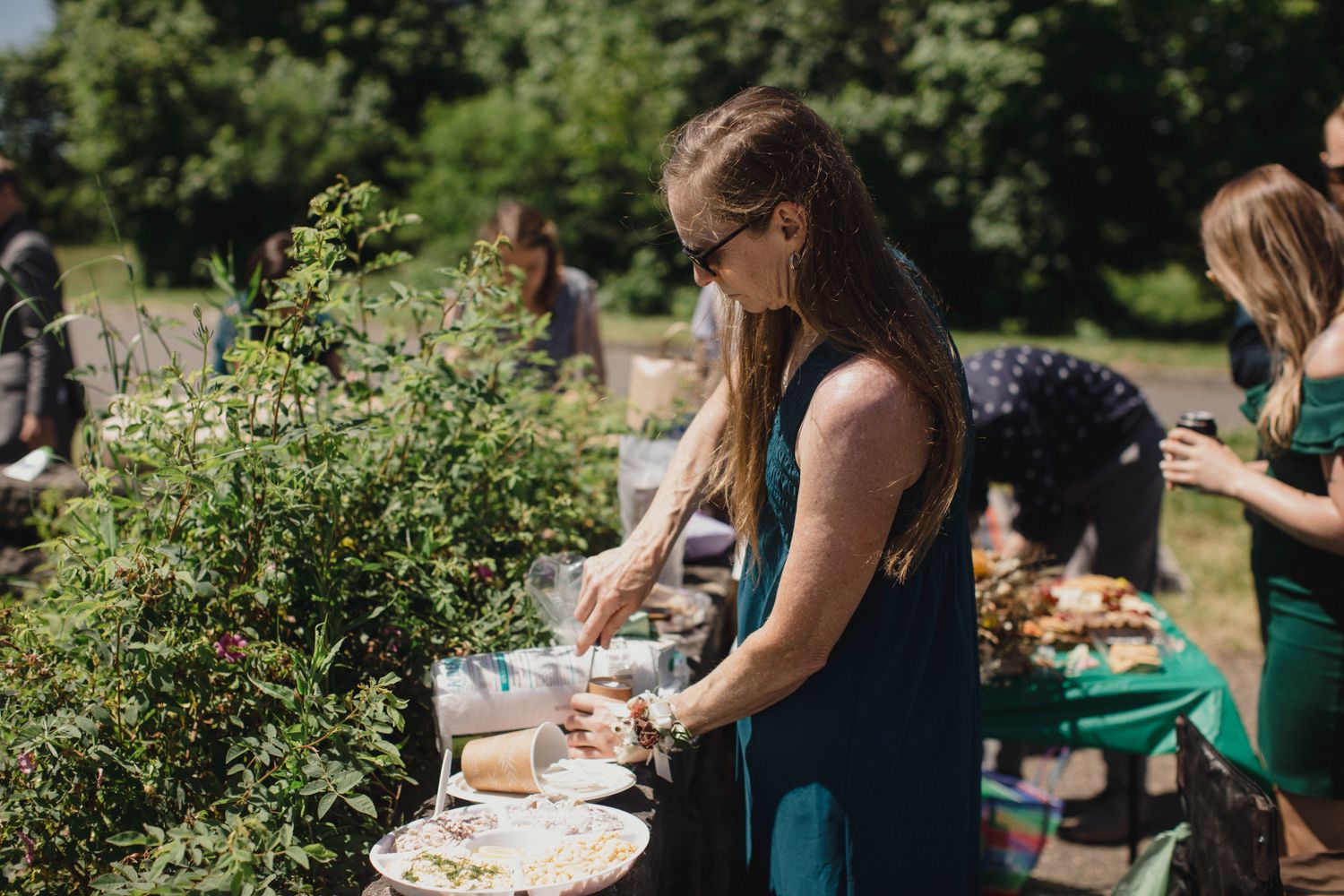 People preparing food at an outdoor buffet table surrounded by greenery.