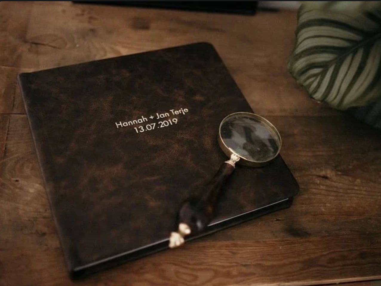 Dark leather wedding guest book for Hannah and Jan Teije dated 13.07.2019 with a vintage magnifying glass on wooden table.