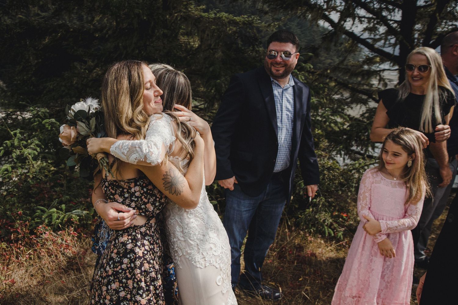 Bride in lace gown shares a warm hug with a guest while family and friends gather outdoors after the ceremony.