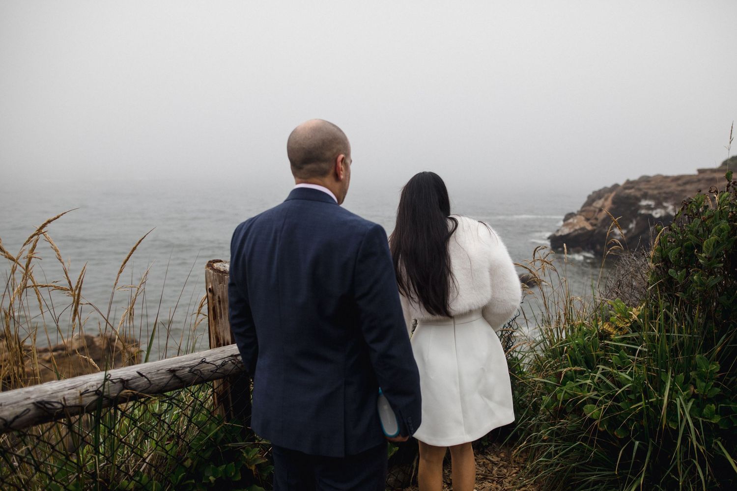 Bride and groom standing at a coastal overlook on a misty foggy wedding day.