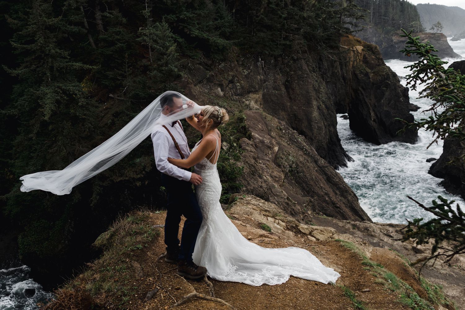 Newlyweds embrace on a cliff edge while the bride's veil flows dramatically in the wind by the ocean.