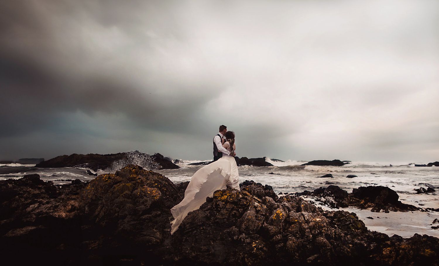 A bride and groom embrace on rocky coastal cliffs with dramatic stormy ocean waves and overcast skies behind them.