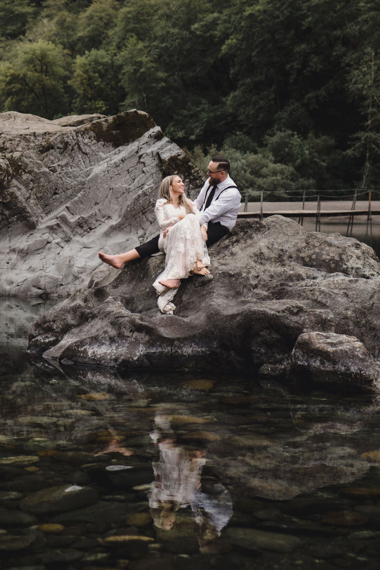 Bride and groom sit together on large rocks beside a reflective mountain lake in wedding attire.
