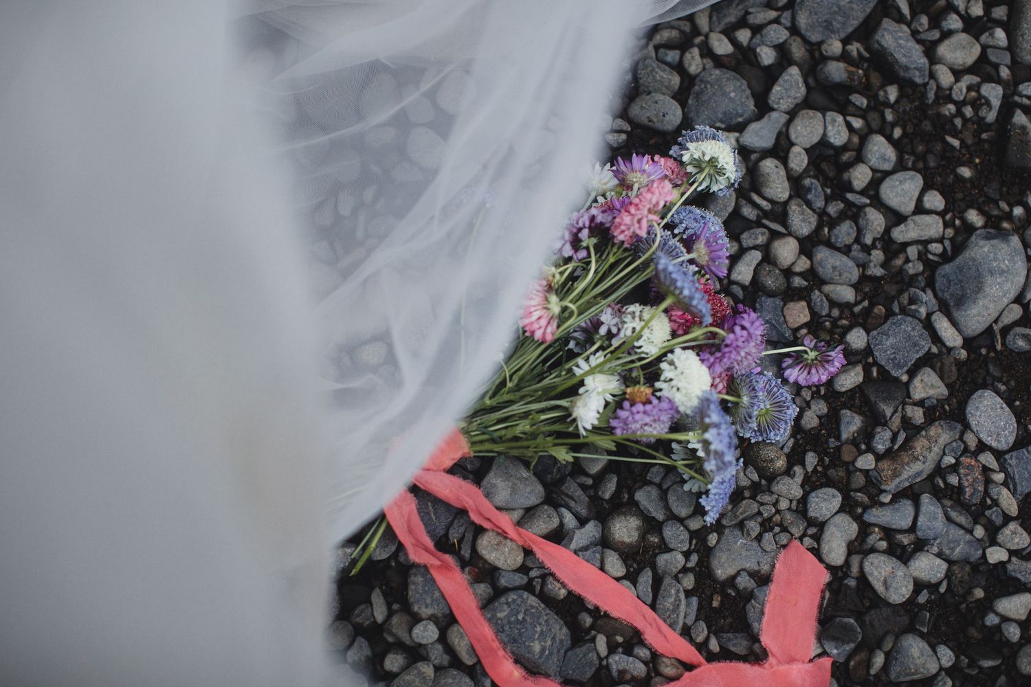Colorful wildflower bouquet with purple and white blooms resting on dark pebbles beside a bridal veil.