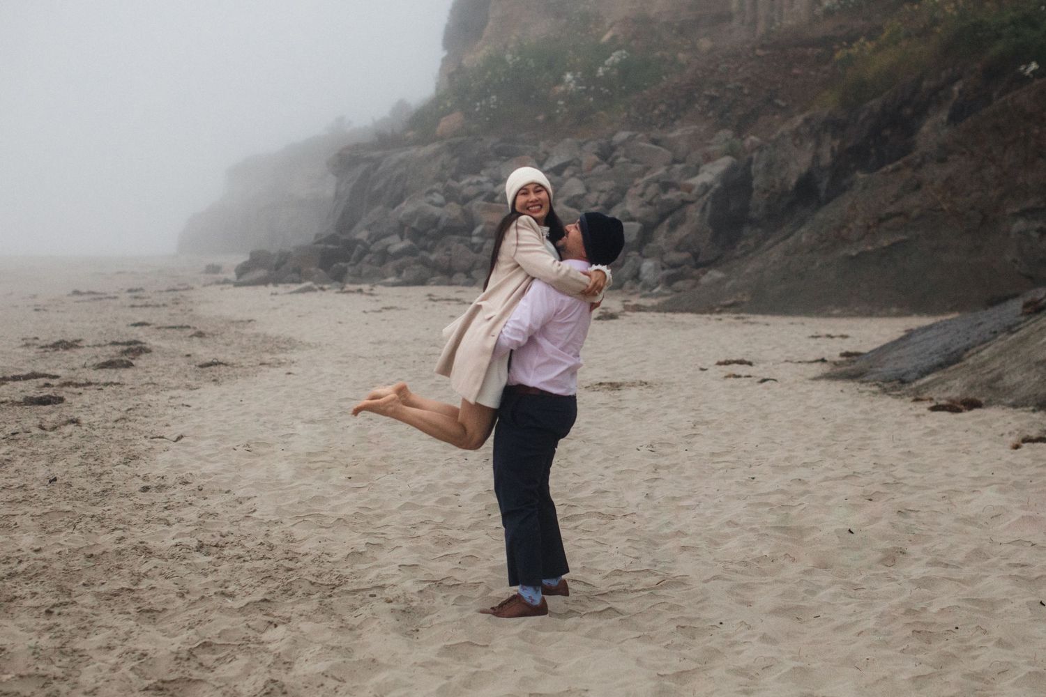 A couple embraces on a misty beach with dramatic cliffs in the background during an overcast day.