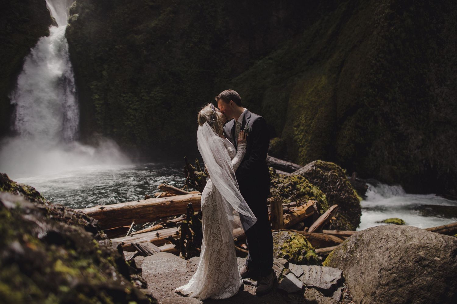 A couple in wedding attire embraces near a misty waterfall and rocky terrain.