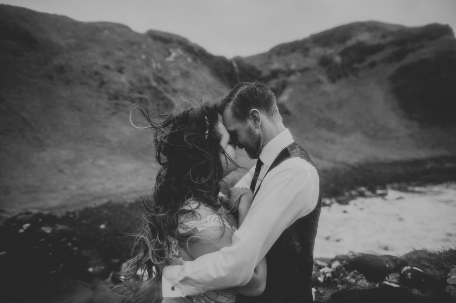 Black and white portrait of couple embracing on windswept coastal landscape.