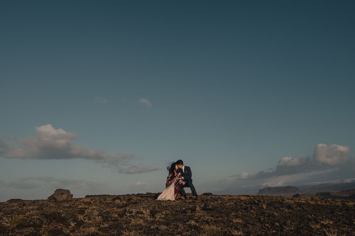 A dramatic wide shot of a couple on rocky terrain against a vast blue sky.