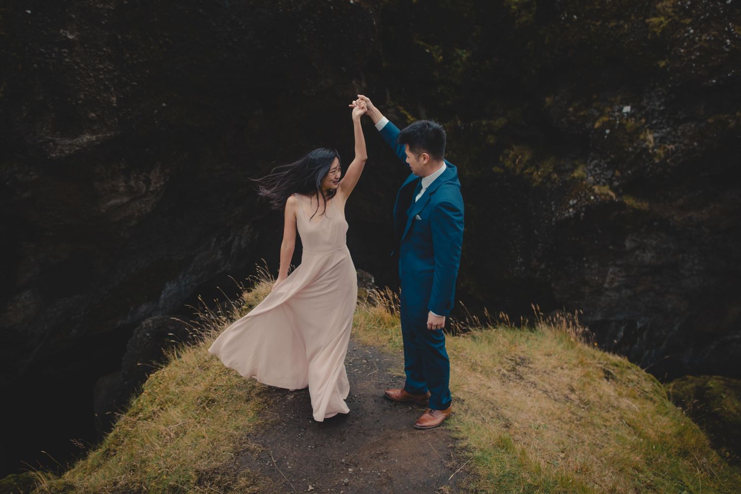 Couple in formal wear dance together on mountain path with dramatic backdrop.