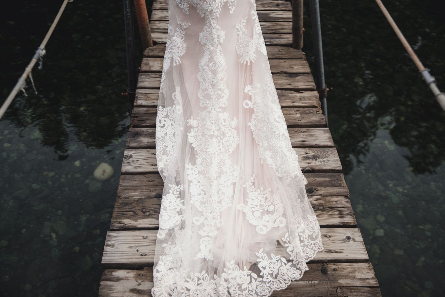 Elegant lace wedding dress with blush pink lining displayed on a rustic wooden dock over dark water.