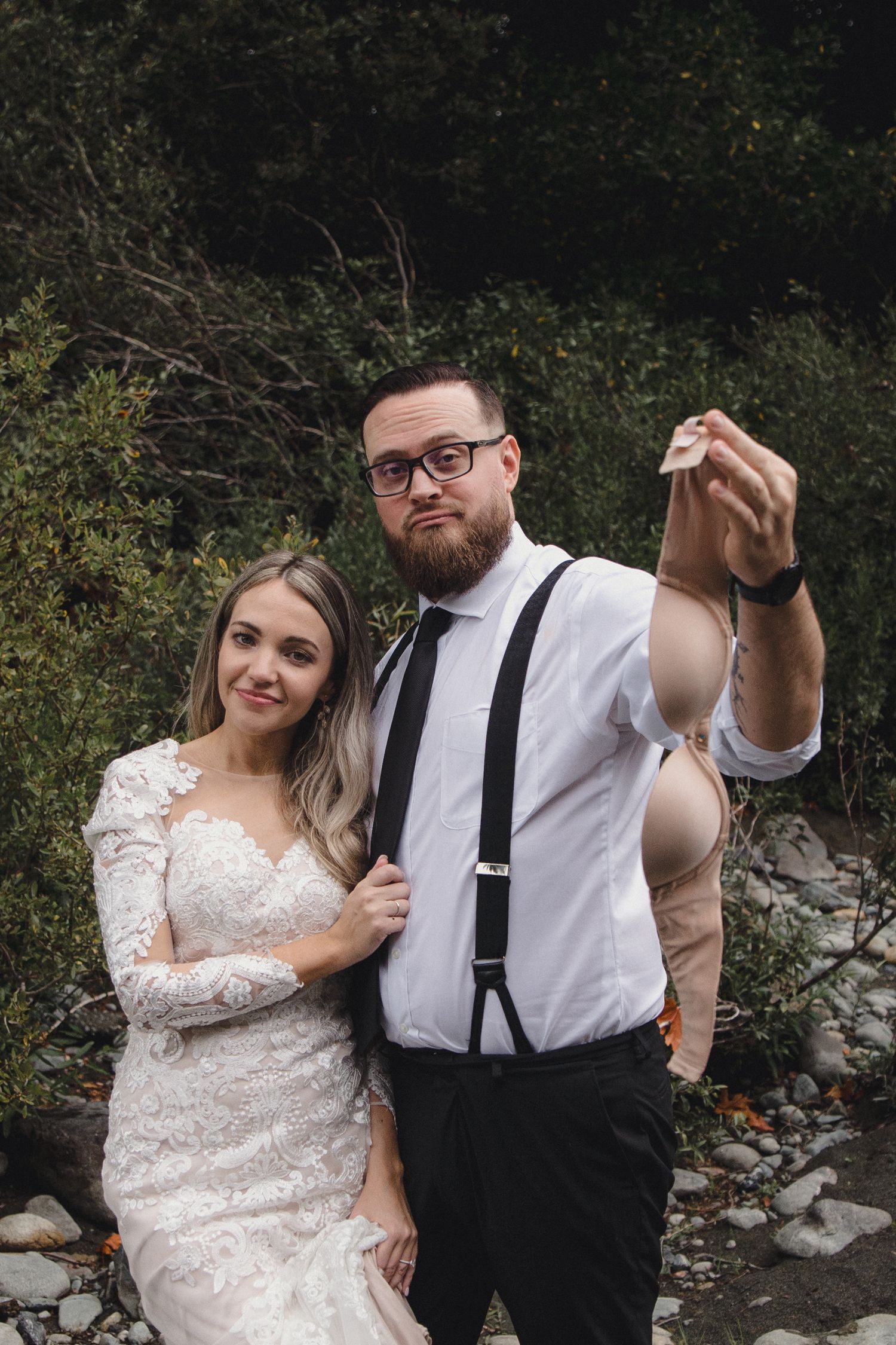 Bride and groom pose outdoors, groom holding up a small ring on a rocky nature backdrop.