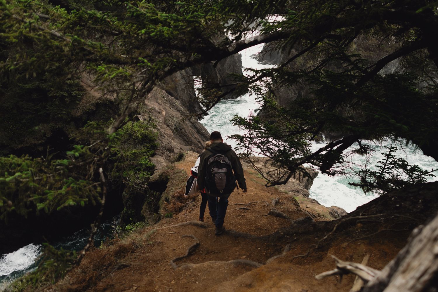 Hiker ascending rugged mountain path alongside snowy terrain and evergreen trees.
