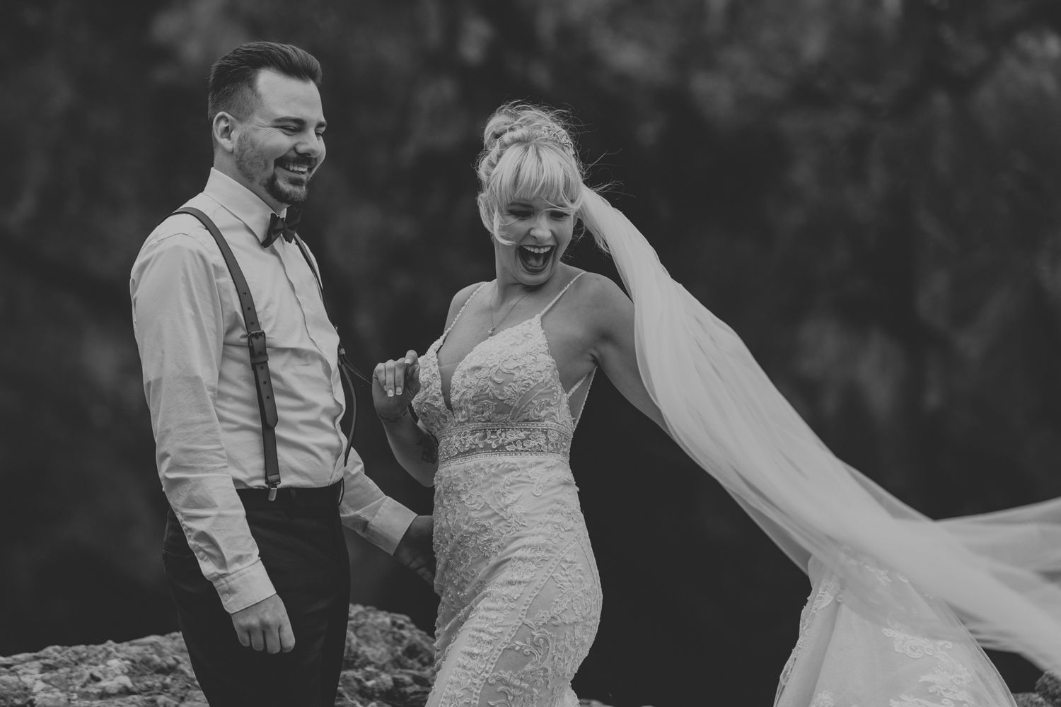 A black and white outdoor wedding photo showing a couple in elegant attire with a flowing veil catching the breeze.