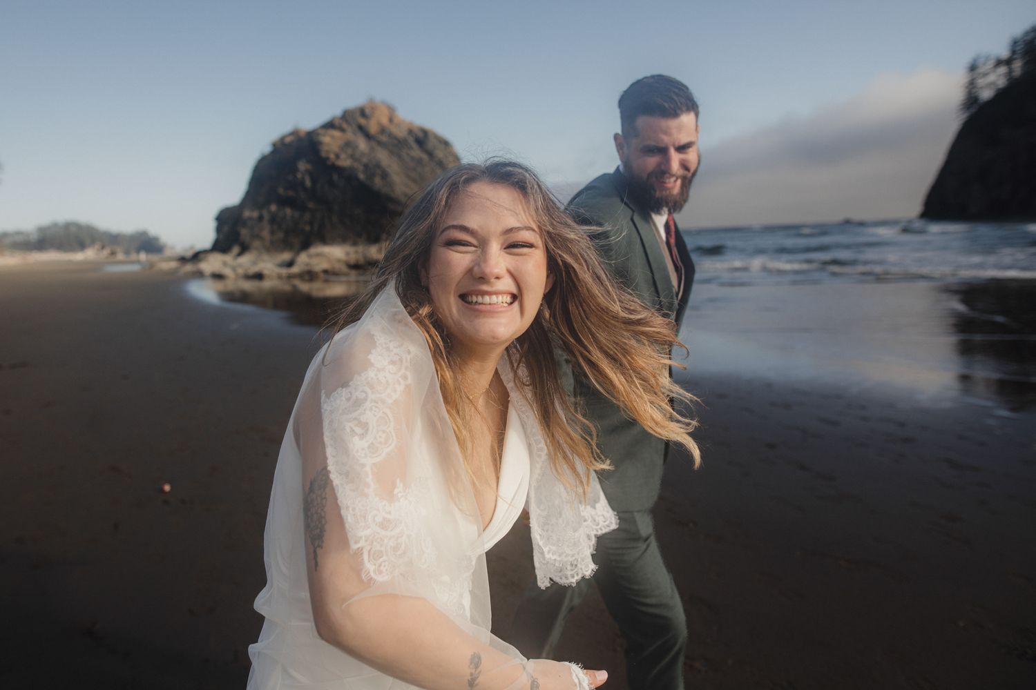 Bride and groom laughing on a black sand beach with ocean waves and rocky cliffs at golden hour.