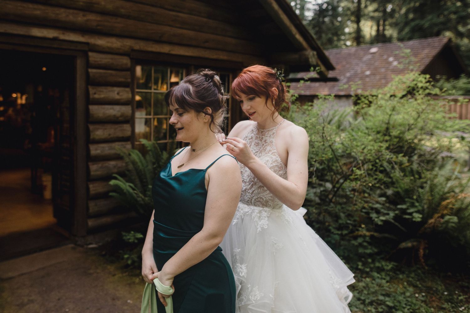 Bridesmaid in emerald green dress helps red-haired bride with her lace wedding gown outside a rustic log cabin.
