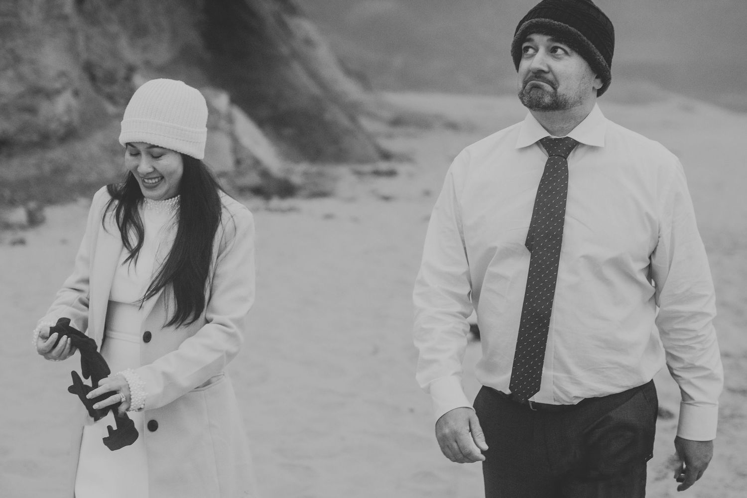 Black and white photo of couple walking on a misty beach in semi-formal attire.