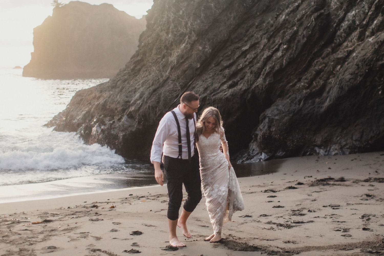 Barefoot bride and groom walk hand in hand along a sandy beach at golden hour near dramatic coastal cliffs.