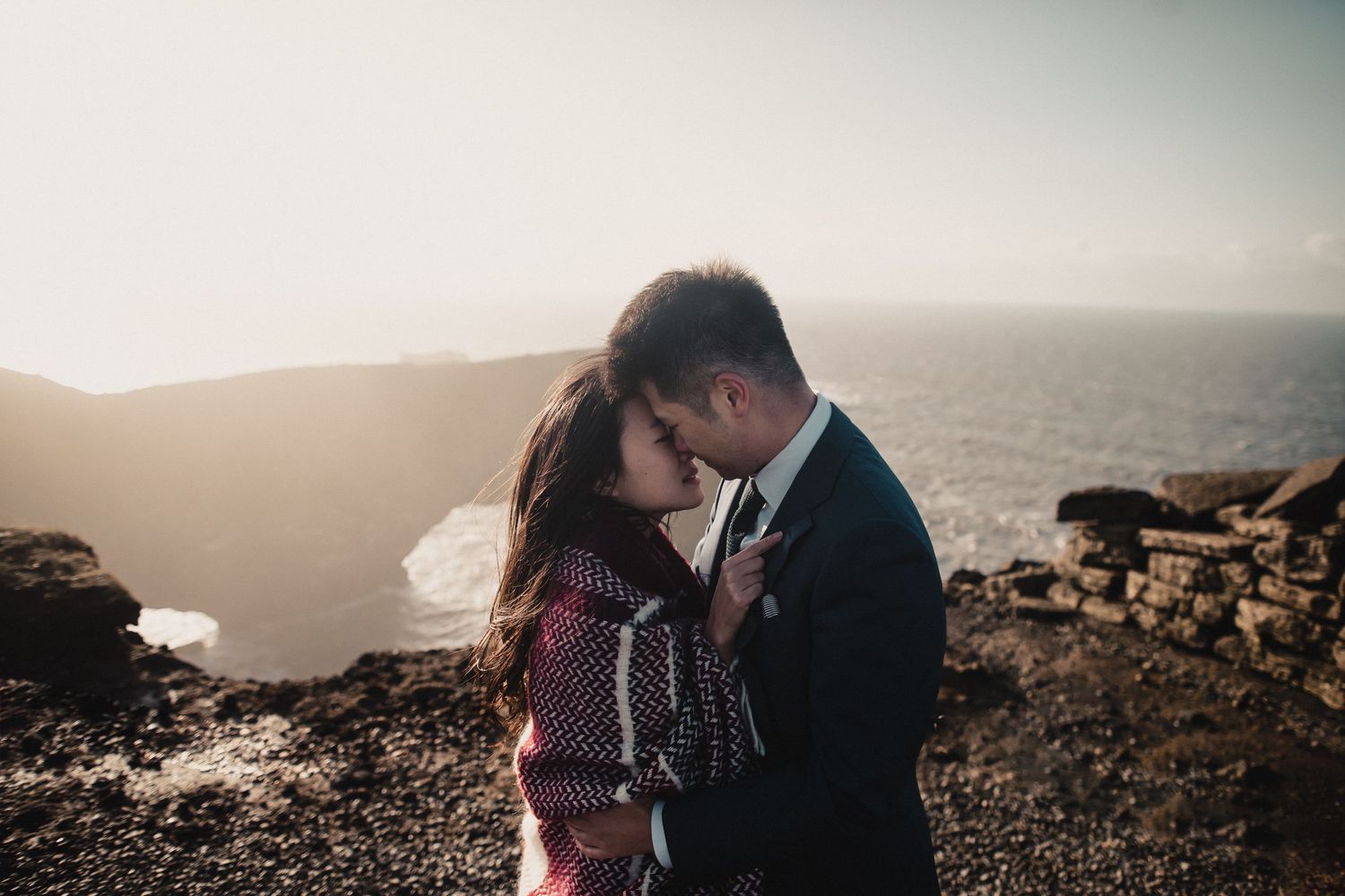 Couple sharing intimate moment on rocky coastal overlook at sunset.