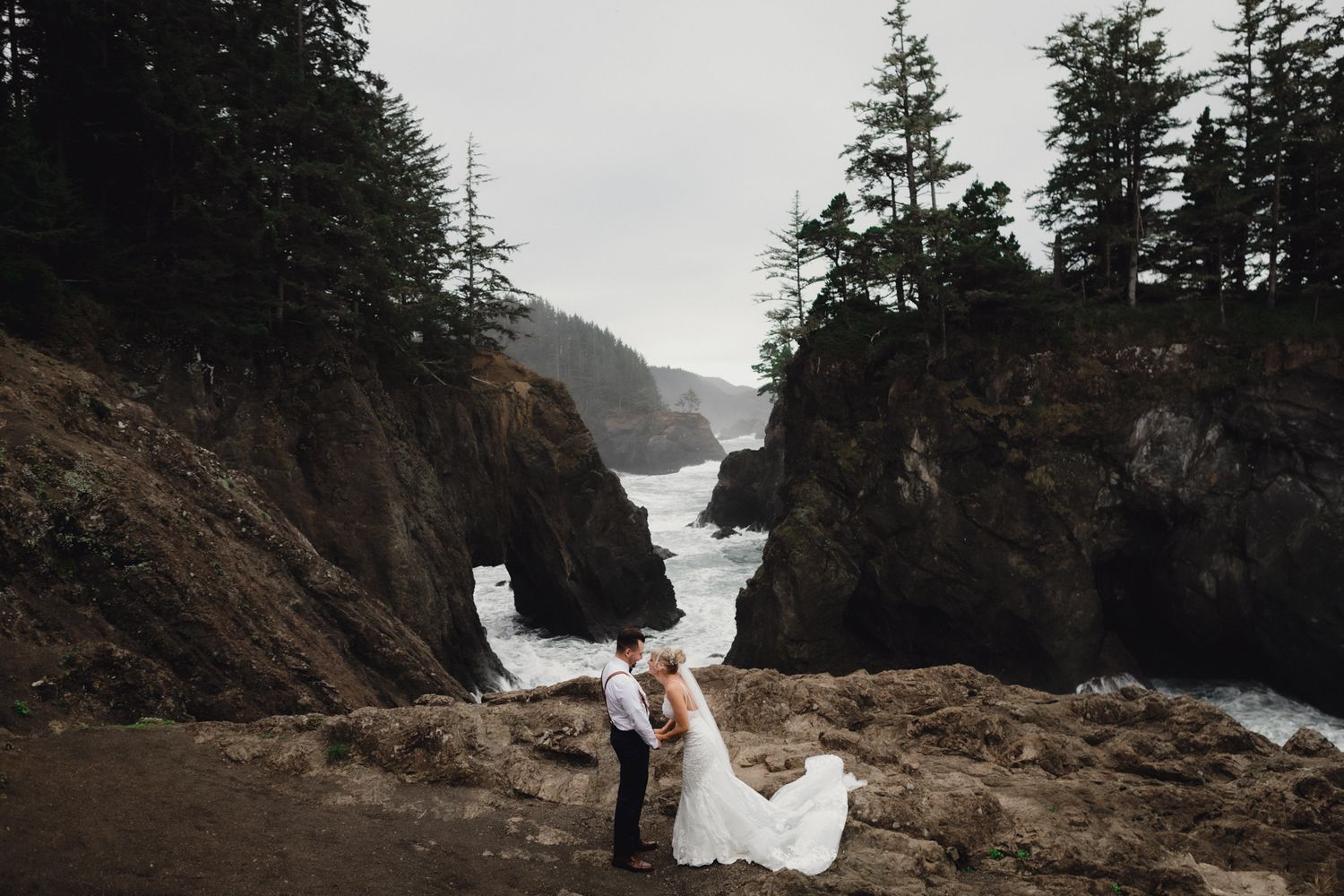 A wedding couple embraces on dramatic coastal cliffs with rugged ocean views and evergreen trees.
