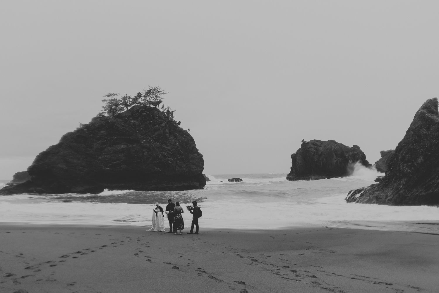 A black and white beach scene shows silhouettes walking among dramatic coastal rock formations.