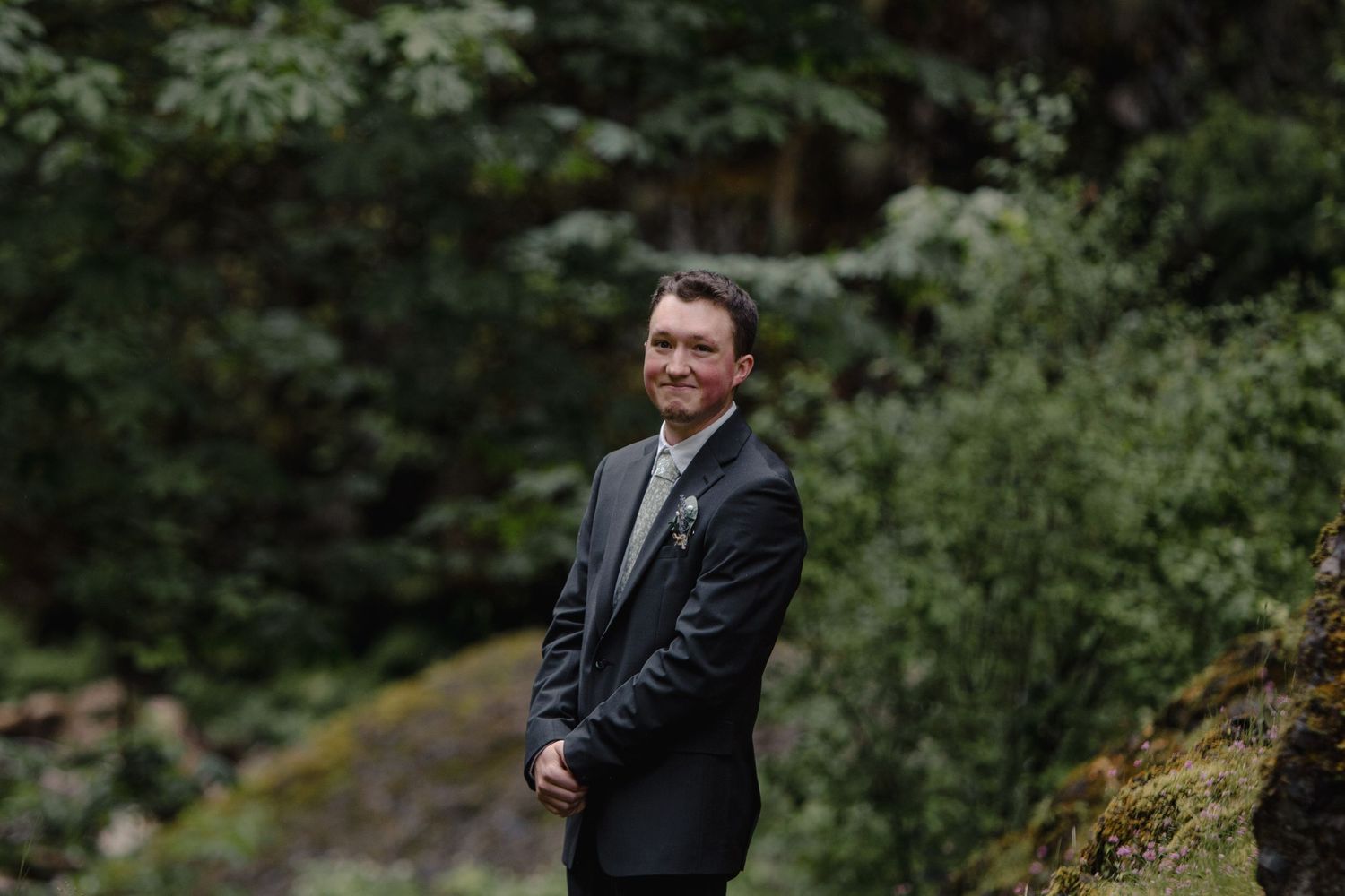 A suited figure smiling while standing on a mossy forest path.