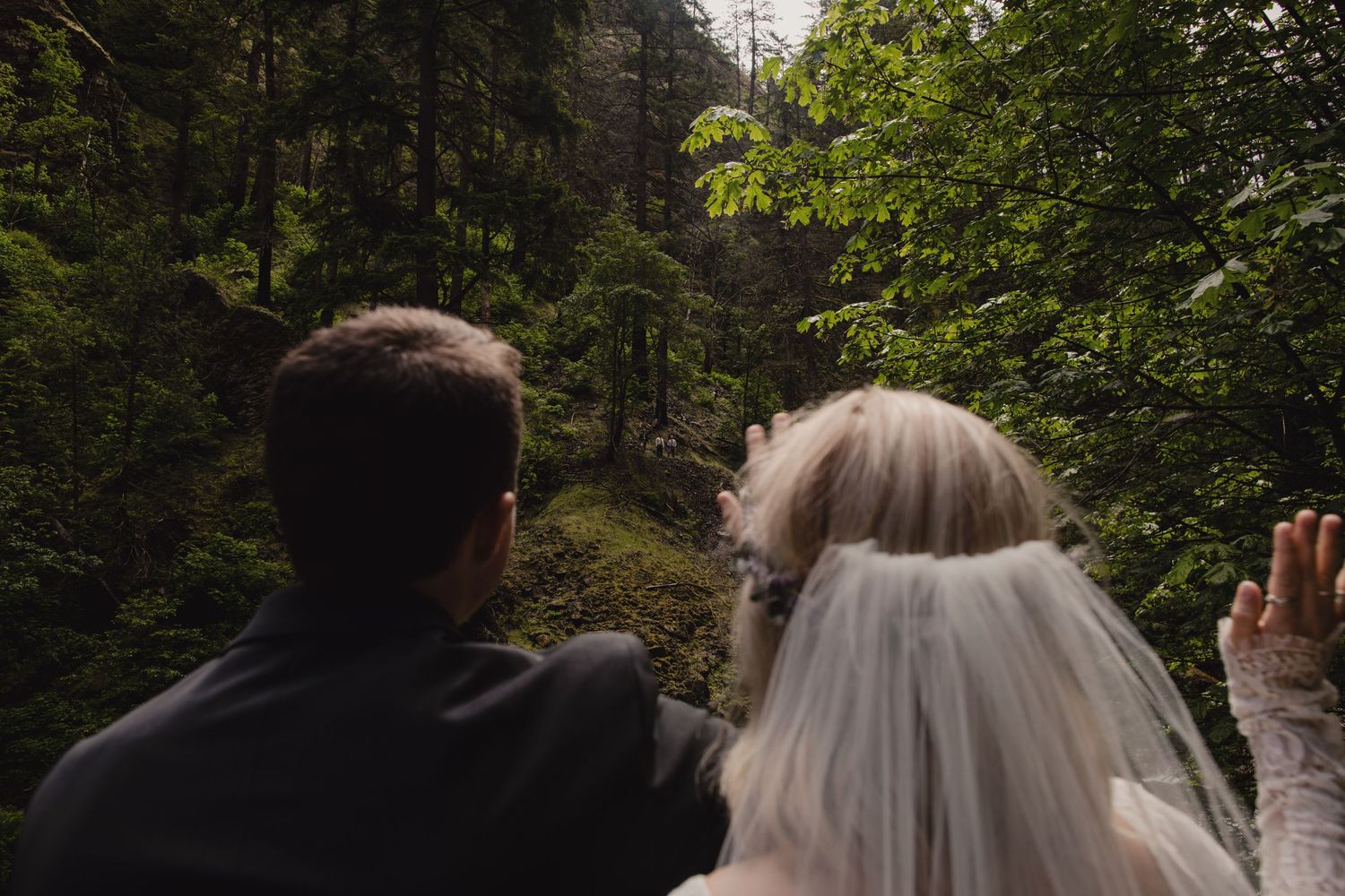Couple in wedding attire views scenery from forest path with green foliage overhead.